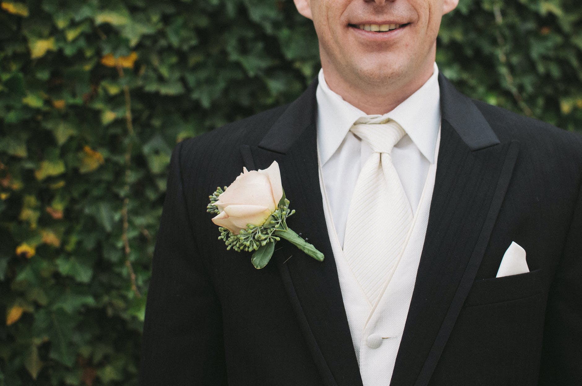 man in black formal suit with white necktie beside green bush in shallow focus photography